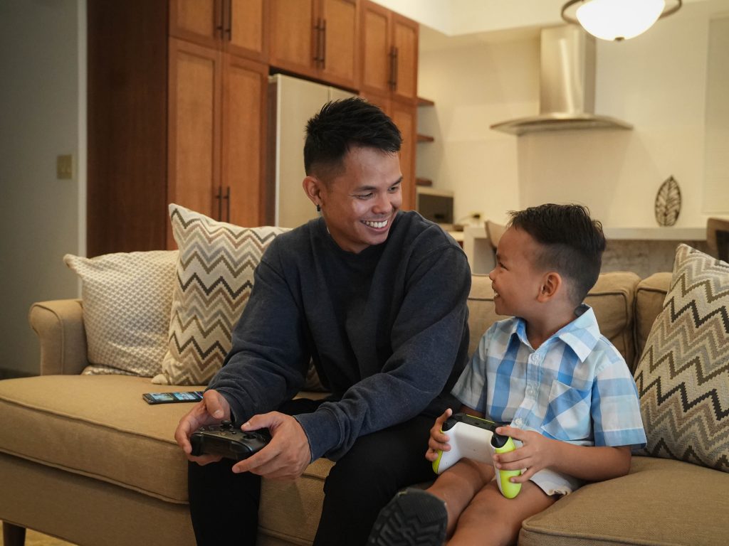  A young father and soon sit on a living room couch with video game controllers in hand. They are looking at each other as the enjoy time spent together playing video games supported by IT&E's Home Internet.